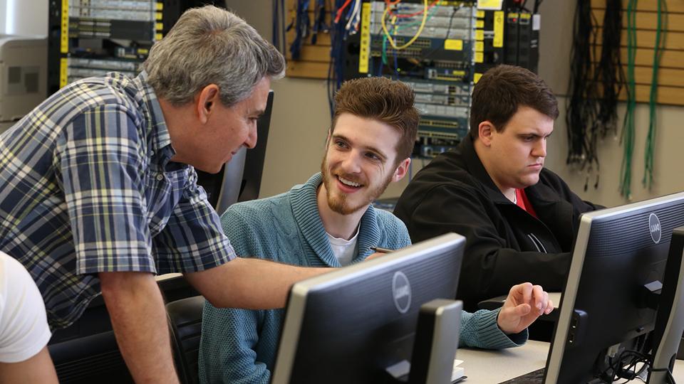 A student talks with his professor in a networking class while another student works on a project.